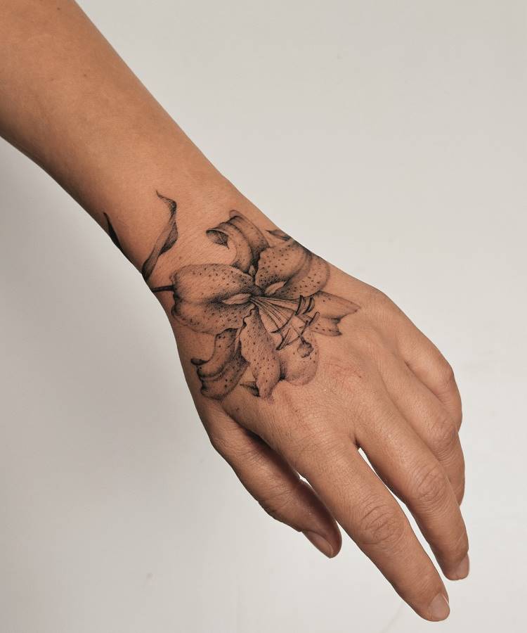 Tatuagem em preto e cinza no dorso da mão, com uma flor de lírio detalhada, linhas finas e sombreamento pontilhado. Os detalhes incluem pétalas delicadas e folhas sutis. Fundo branco simples, destacando a pele e a arte minimalista.