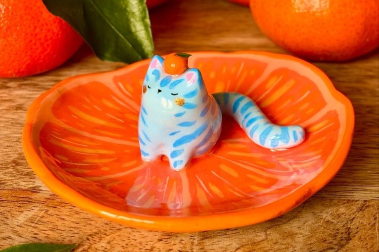 Miniatura de cerâmica fria com forma de gato azul listrado, sentado em um prato laranja com textura de fatia de fruta. O gato tem um pequeno tangerina na cabeça e está rodeado por laranjas naturais decorativas.