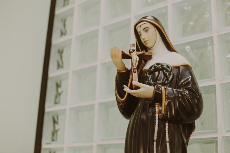 Imagem de uma estátua de Santa Rita de Cássia segurando um crucifixo com expressão serena, em frente a um fundo de azulejos claros.