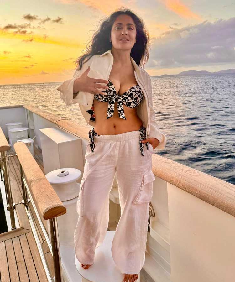 Salma Hayek Mulher de cabelos soltos e ondulados posa em um iate ao pôr do sol. Ela usa calça branca, blusa aberta e top estampado. O céu colorido e o mar azul criam uma atmosfera tropical e tranquila.