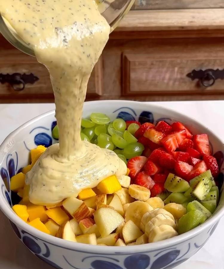 Molho cremoso é derramado sobre uma variedade colorida de frutas em uma tigela grande. A mistura inclui pedaços de maçã, manga, kiwi, uvas, bananas e morangos. O cenário é uma cozinha rústica com armários de madeira ao fundo.