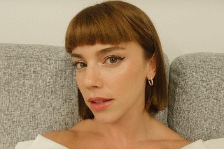Mulher com cabelo castanho curto, franja reta, maquiagem leve com delineado nos olhos e batom nude. Usa brincos prateados e está sentada em um sofá cinza claro, olhando diretamente para a câmera.