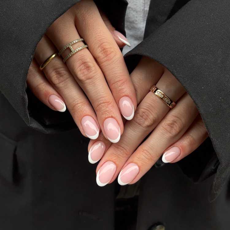 Mão feminina com unhas pintadas de esmalte rosa claro, segurando levemente a perna, vestindo calça bege e tênis branco com listras marrons. Detalhes incluem relógio dourado no pulso e casaco felpudo branco, compondo visual casual e elegante.