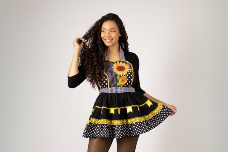 Mulher sorridente com cabelos cacheados escuros, vestindo um avental preto de festa junina. O avental tem estampa de poá branco, detalhes amarelos, uma faixa xadrez azul e um aplique de girassol e flores coloridas. Ela segura a barra da saia do avental com uma mão. Fontes