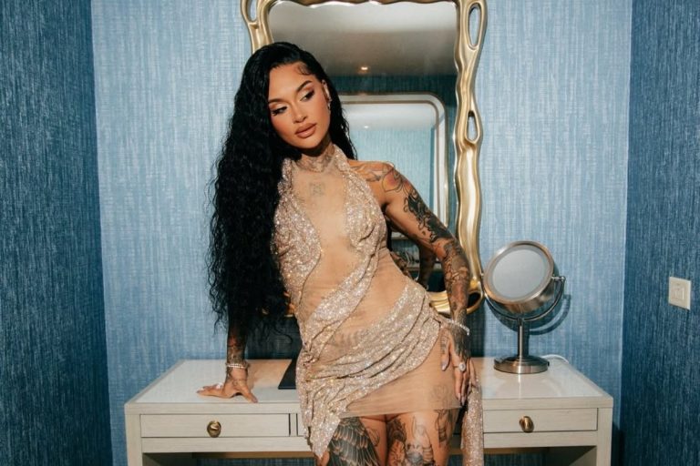Kehlani, com cabelos longos e escuros, posa em um vestido dourado brilhante com detalhes transparentes. Ela está em um quarto com parede texturizada azul e móveis brancos, exibindo suas tatuagens nos braços e pernas
