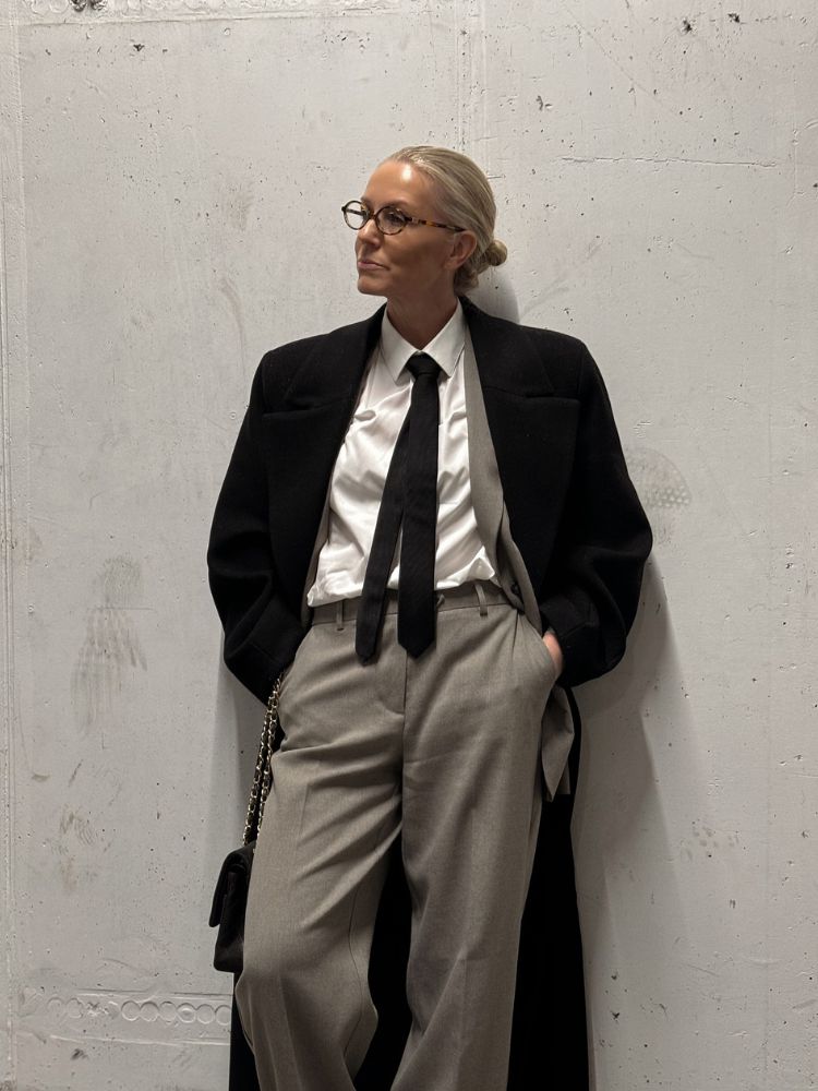 Senhora de pele clara de camisa branca, gravata preta, blazer preto e calça alfaiataria cinza