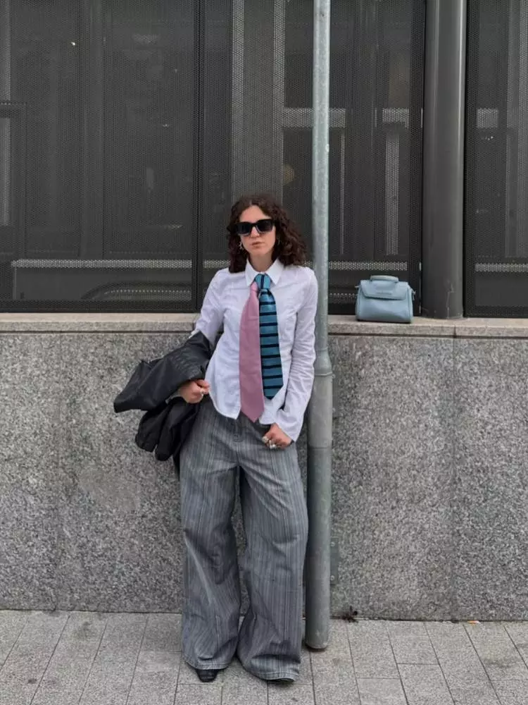 Mulher de pele clara de camisa branca, gravatas azul e rosa, calça jeans, segurando jaqueta preta