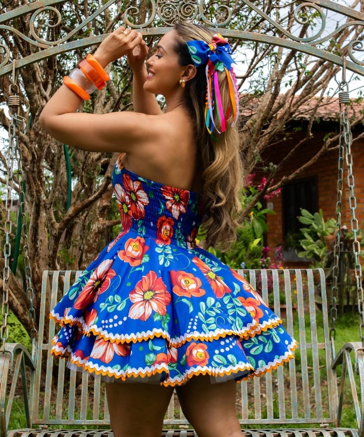Uma mulher sorridente, de pele clara e cabelos castanhos ondulados, está em um balanço de jardim. Ela veste um vestido azul vibrante com estampas florais laranjas e brancas, detalhes em babados e uma faixa de cabelo azul com fitas coloridas. Ela usa pulseiras laranjas e brincos dourados, com as mãos perto do rosto em um gesto alegre. Ao fundo, árvores e uma casa de tijolos sugerem um ambiente festivo ao ar livre, possivelmente no Brasil, dada a vestimenta que lembra trajes típicos de festas juninas.