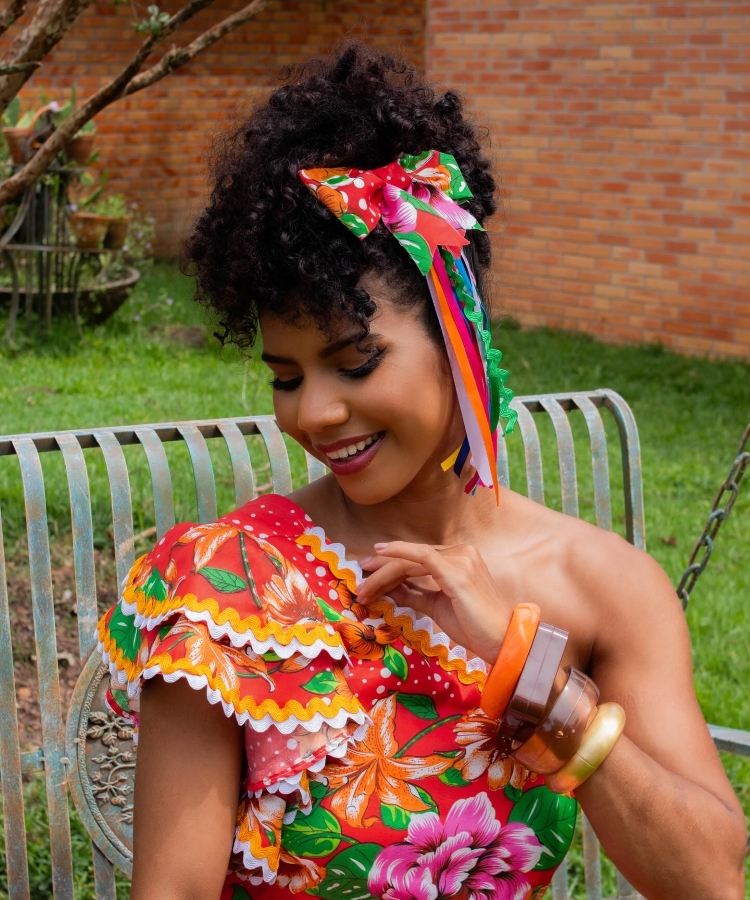 Uma mulher sorridente de pele morena clara está sentada em um banco de metal em um jardim. Ela veste um vestido colorido com estampas florais e detalhes em renda branca e amarela. Cabelos escuros e cacheados estão adornados com um laço estampado e fitas coloridas. Ela usa pulseiras laranjas, marrons e douradas em um dos braços. Ao fundo, vê-se grama verde e uma parede de tijolos.