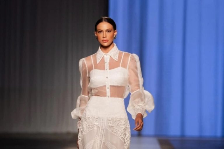 Uma modelo esguia caminha por uma passarela de moda, vestindo uma blusa branca de manga bufante e transparente sobre um top tomara que caia, combinando com uma saia longa branca de renda.