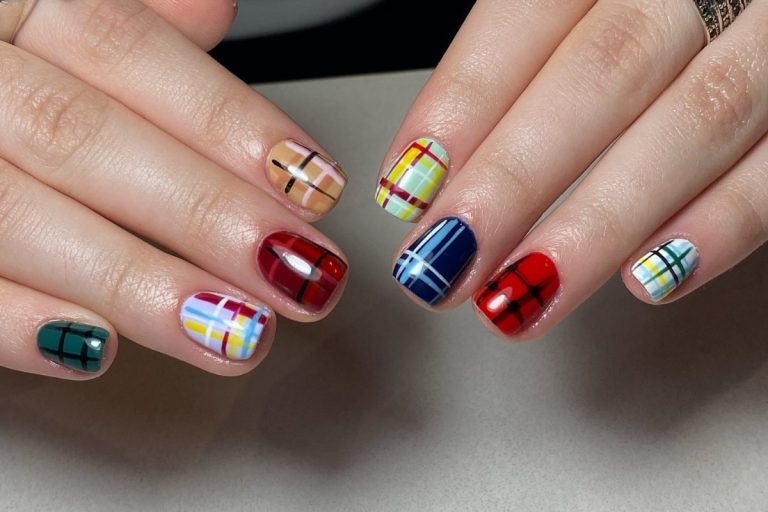 Unhas curtas com esmalte em design xadrez vibrante. Cada unha apresenta uma cor de base diferente (verde, roxo claro, vermelho, azul, vermelho, amarelo claro, bege claro) com listras pretas e brancas cruzadas, criando um efeito de padrão de tartan. Fontes