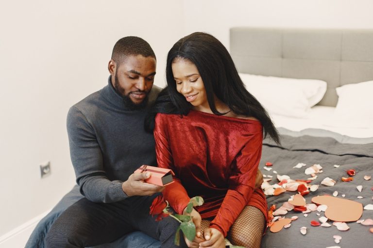 Homem de pele negra entregando presente para mulher de pele negra na cama enfeitada com flores