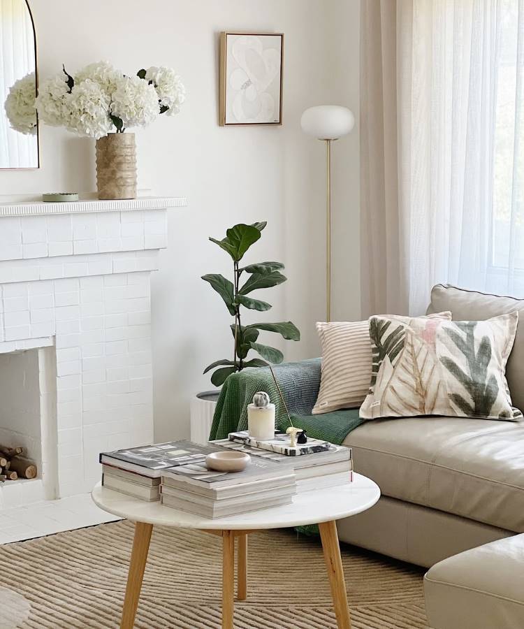 Uma sala de estar clara com sofá bege, almofadas estampadas e manta verde. Uma mesa de centro redonda exibe livros e decoração. Ao fundo, uma lareira branca, vaso com flores brancas e um espelho. Uma planta alta e uma luminária de piso completam o ambiente.