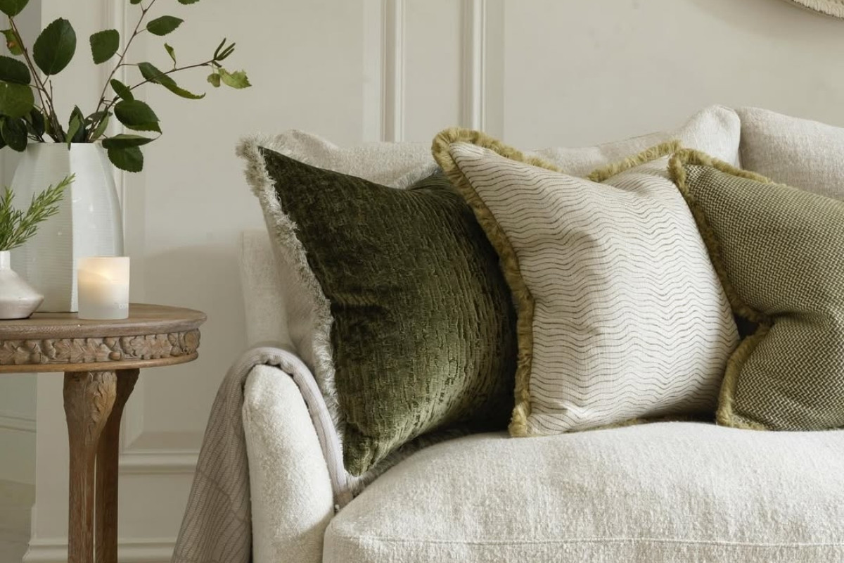 Uma sala elegante com sofá claro e almofadas para sala em tons de verde e creme, com texturas e franjas. Ao lado, uma mesa de madeira redonda exibe um vaso com folhagens e uma vela acesa. Um espelho redondo na parede e uma luminária preta completam o ambiente sofisticado.