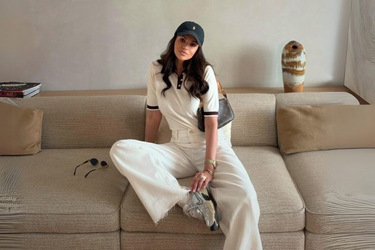Uma mulher sentada em um sofá bege, usando um polo creme, calça jeans branca larga, boné de beisebol preto, óculos de sol, tênis prateados e uma bolsa de ombro marrom.