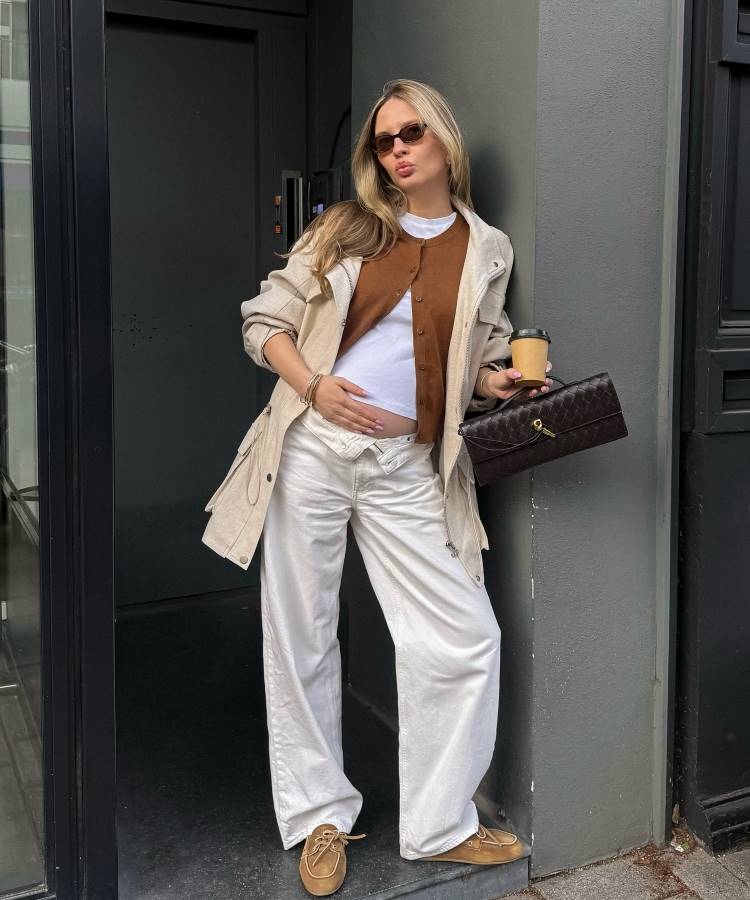 Uma mulher grávida com cabelo loiro comprido posa ao ar livre, vestindo uma camiseta branca, um cardigan marrom, um casaco bege, calças largas prateadas e mocassins marrons, segurando uma bebida e uma bolsa escura. Ela usa óculos de sol e parece estar em um ambiente urbano.