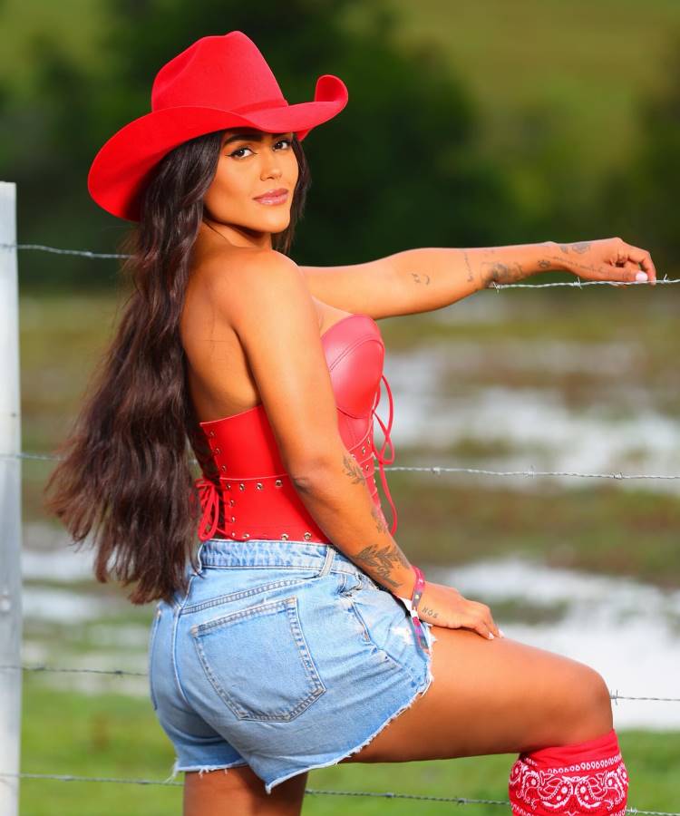 Camila Loures posa com um chapéu de cowboy vermelho, corpete vermelho e shorts jeans. Ela se apoia em uma cerca de arame farpado, exibindo tatuagens nos braços e meias estampadas. O fundo desfocado sugere um cenário rural.