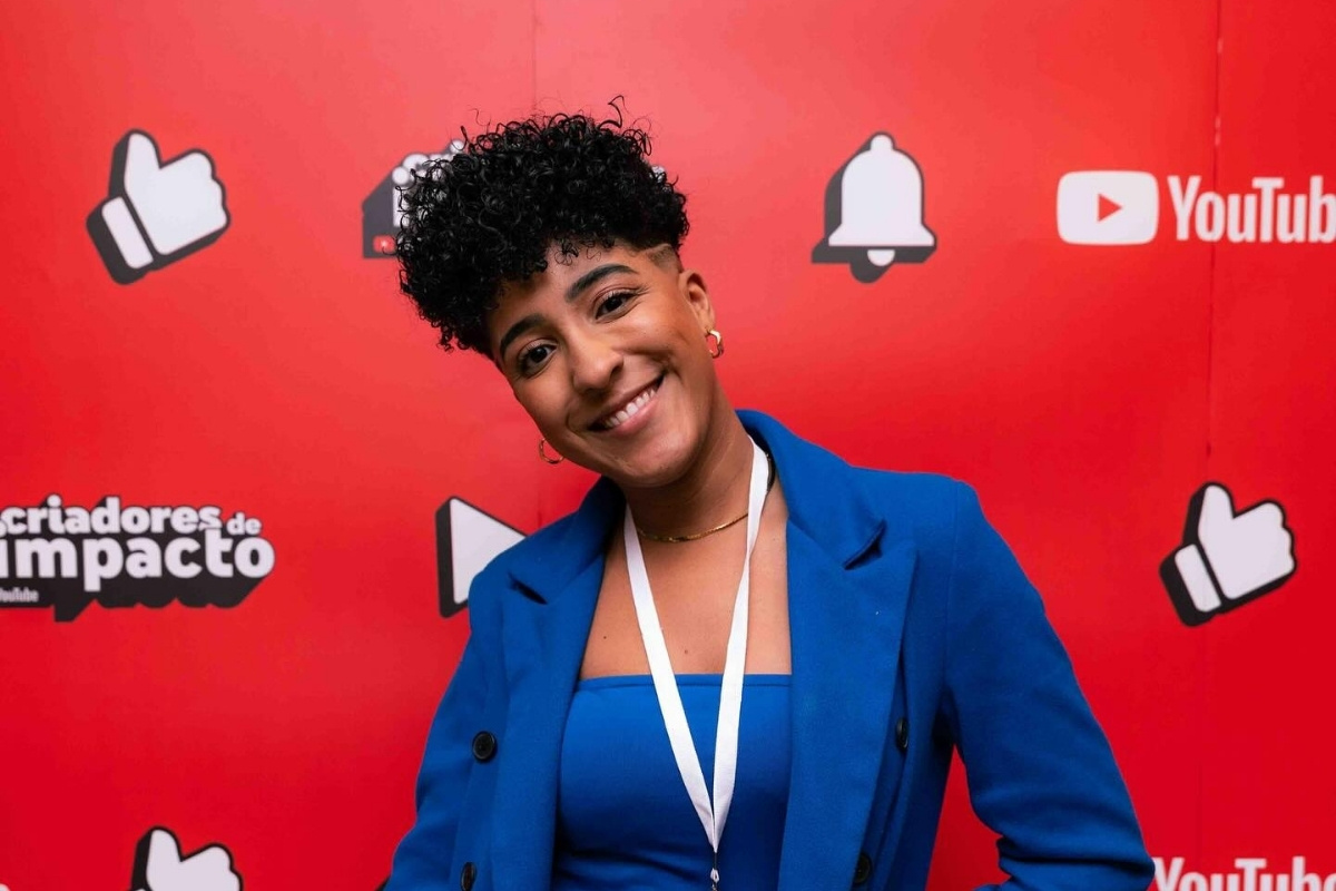 Nath Finanças usando top azul, blazer azul, em frente a um painel do YouTube