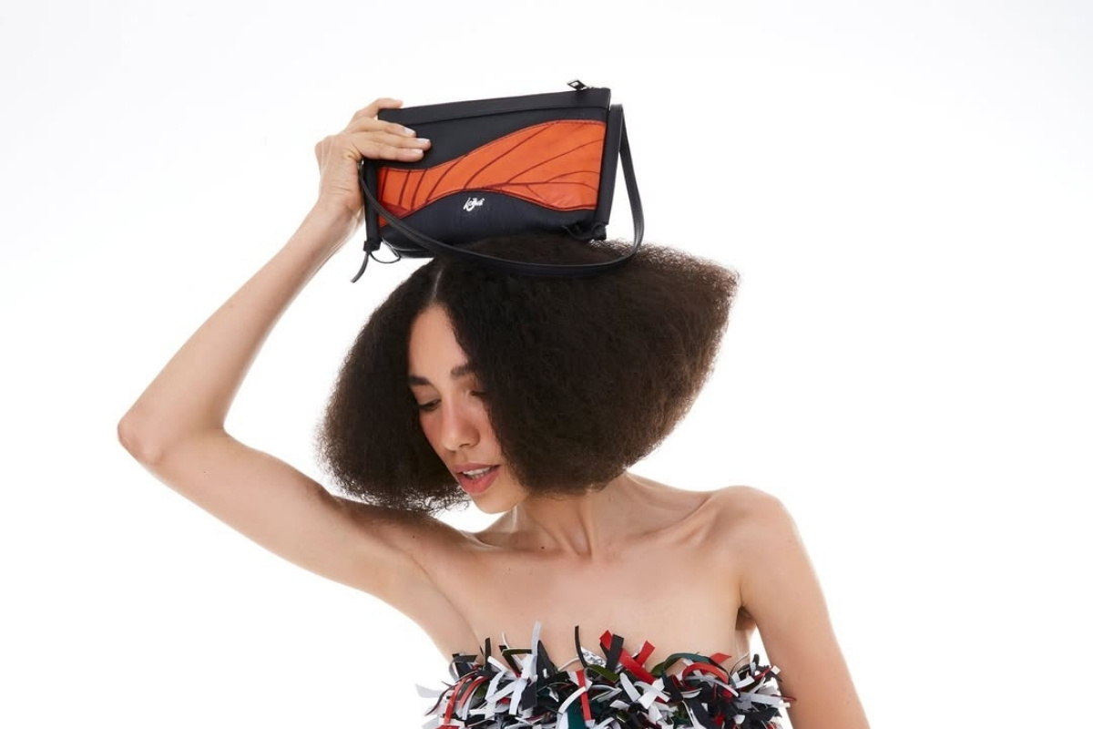 Uma mulher de pele clara e cabelo escuro, com um volume exagerado no cabelo, segura uma bolsa preta e laranja acima da cabeça. Ela usa um top tomara que caia com franjas coloridas. O fundo é branco.