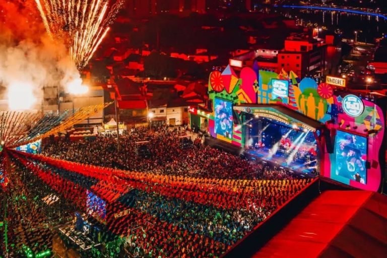 Uma festa junina vibrante à noite, com milhares de pessoas reunidas sob luzes e bandeirinhas coloridas. Fogos de artifício iluminam o céu acima de um grande palco onde artistas se apresentam, cercado por edifícios e casas.