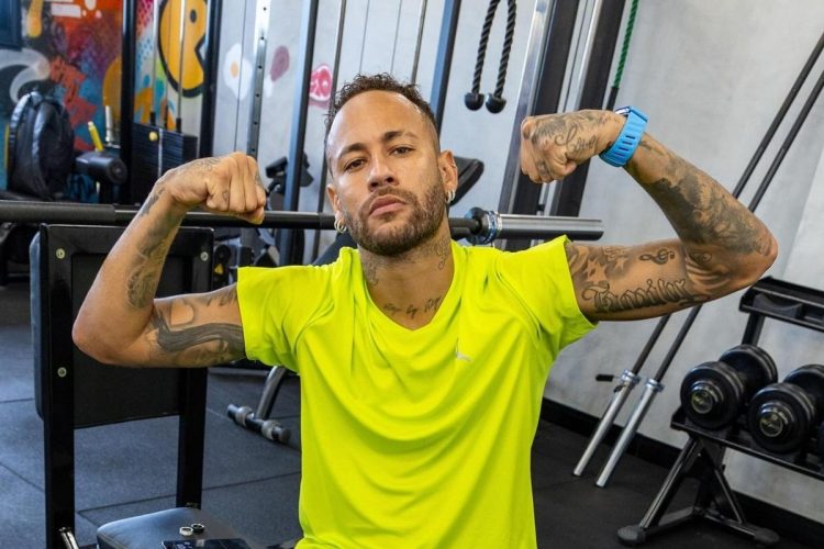 Neymar Jr. posa em uma academia, flexionando os bíceps para a câmera. Ele veste uma camiseta verde-limão e um relógio azul, exibindo suas tatuagens nos braços. O ambiente inclui pesos e equipamentos de ginástica ao fundo.
