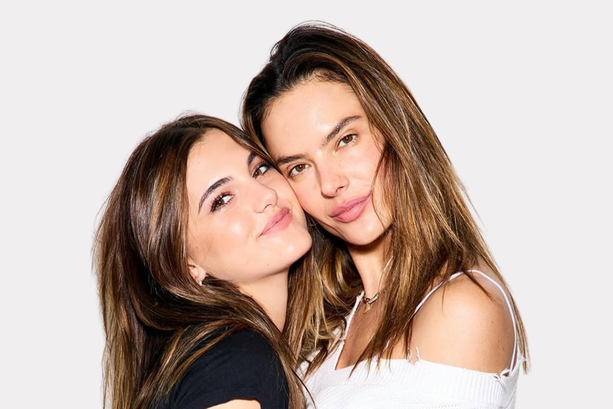 Duas mulheres de cabelos castanhos longos se abraçam. Uma veste camiseta preta e jeans, a outra um suéter branco de ombro a ombro e jeans. Nomes diferentes de filhos de famosos: elas se encostam, sorrindo gentilmente para a câmera, transmitindo uma conexão calorosa.