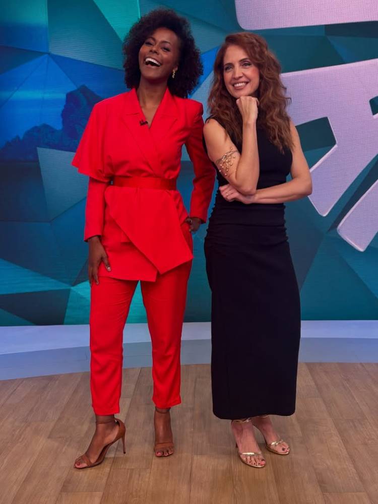 Duas mulheres posam em um estúdio. Maju Coutinho, com cabelo encaracolado e terno vermelho, sorri amplamente. A outra, com cabelo ondulado e vestido preto, tem a mão no queixo e um bracelete dourado no braço.