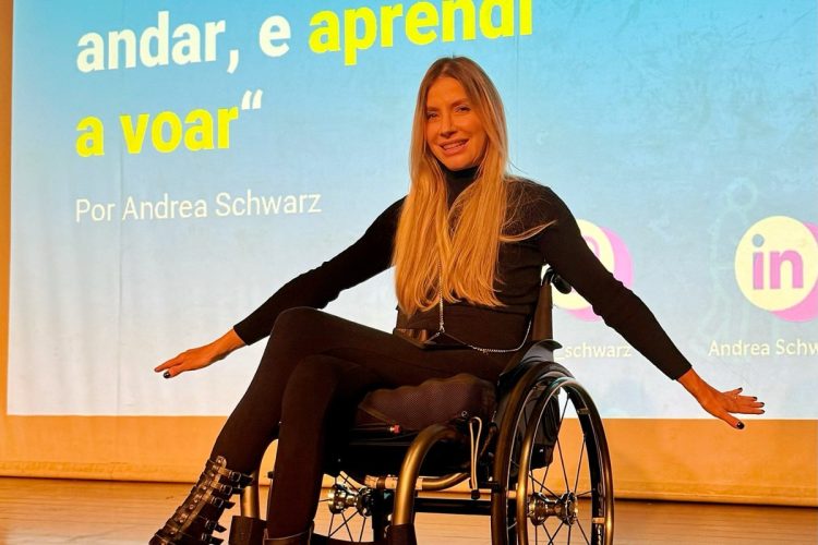 Andrea Schwarz de look preto com painel azul ao fundo escrito "Eu Parei de Andar e Aprendi a Voar" e suas redes sociais