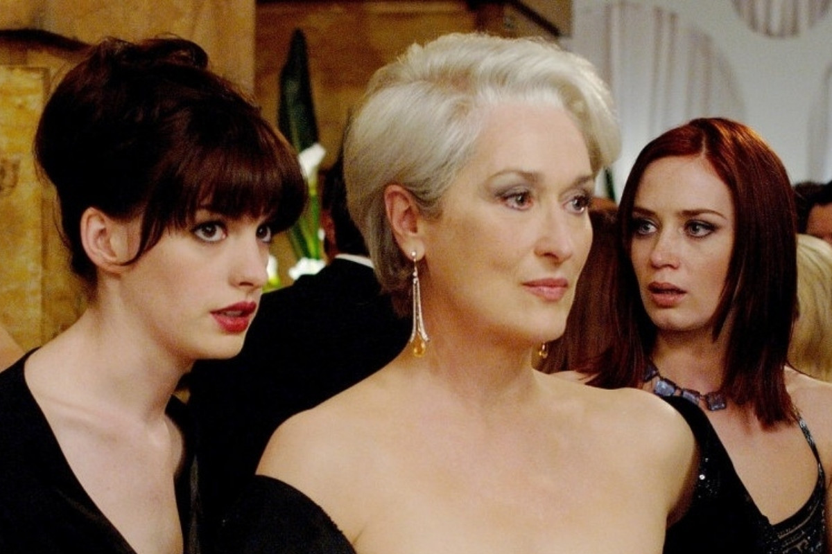 A imagem mostra três atrizes do filme "O Diabo Veste Prada". Meryl Streep, com cabelo grisalho e brincos longos, está ao centro. À esquerda, Anne Hathaway tem cabelo escuro preso. À direita, Emily Blunt tem cabelo ruivo.