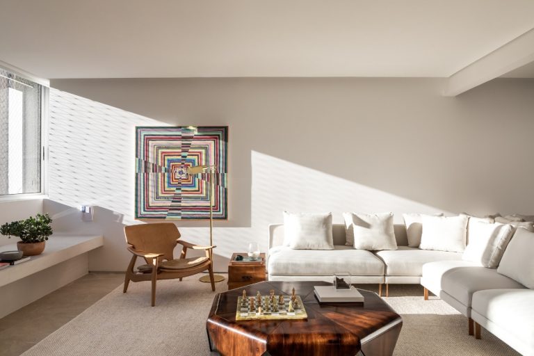 A imagem exibe uma sala de estar clara e contemporânea. Um sofá branco e uma poltrona de madeira clara são o destaque, enquanto uma vibrante obra de arte abstrata adorna a parede. A luz natural cria padrões na parede, e um tabuleiro de xadrez repousa sobre a mesa de centro de madeira.