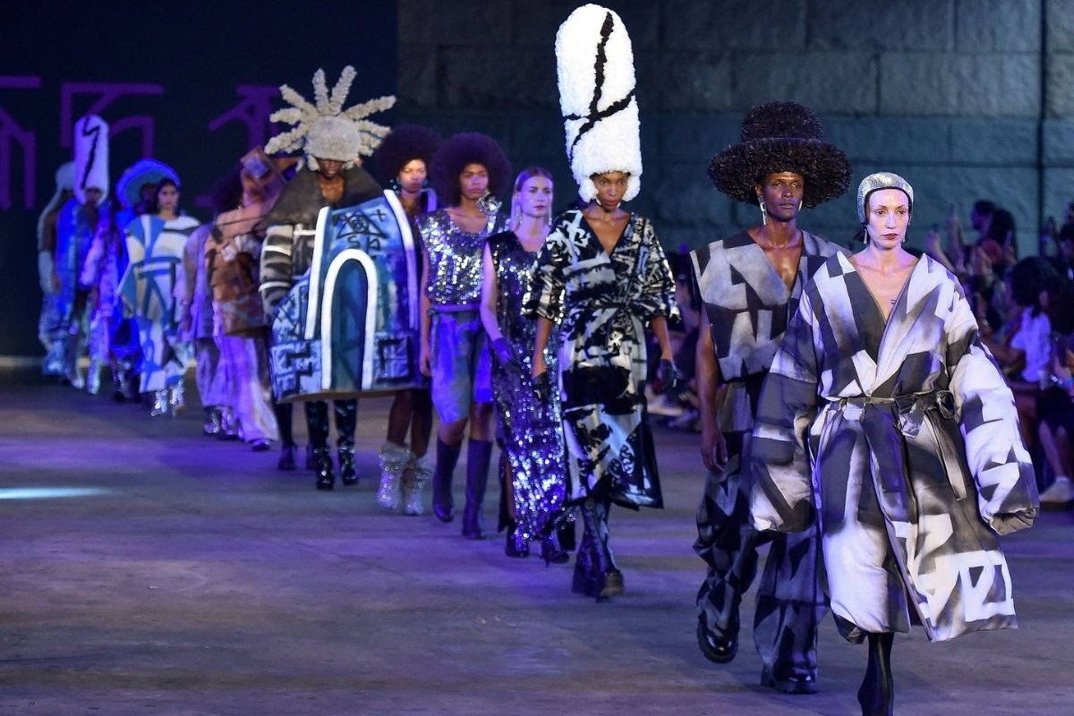 A imagem mostra um desfile de moda da Casa de Criadores, a edição 55, com modelos caminhando em uma passarela ao ar livre. Os modelos exibem uma variedade de roupas e estilos, incluindo: Roupas oversized e com formas geométricas: Muitos dos modelos vestem casacos ou túnicas volumosas com padrões gráficos em preto, branco e cinza. Adereços de cabeça elaborados: Vários modelos usam chapéus grandes e esculturais, alguns em tons neutros como branco e preto, e outros com designs mais exóticos, como o que se assemelha a raios. Texturas e brilhos: Há modelos com roupas mais justas e brilhantes, algumas com lantejoulas ou tecidos metálicos. Diversidade de modelos: Há modelos de diferentes etnias e tipos de cabelo, incluindo afros volumosos. A iluminação sugere um evento noturno, com a passarela iluminada e um público visível ao fundo. O ambiente parece ser em uma área urbana ou um espaço aberto, com construções ao fundo.