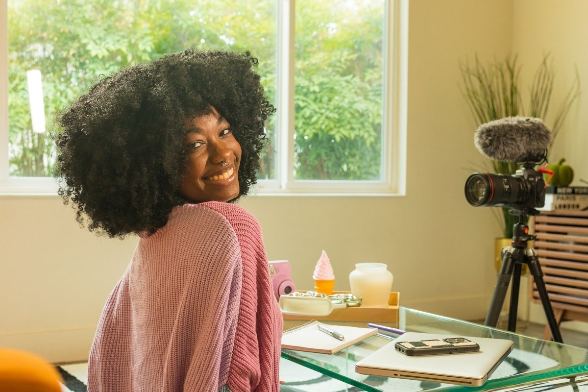 Uma jovem sorridente, com cabelo crespo volumoso, olha para trás em direção à câmera, com um microfone e câmera profissional em tripé ao lado. Ela está em um ambiente claro, que remete a um estúdio de criação de conteúdo, com uma mesa de vidro e objetos decorativos.