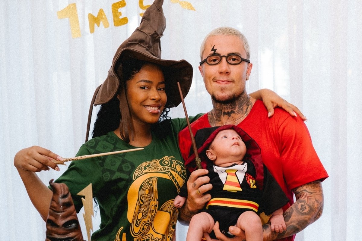 Uma família vestida com fantasias de Harry Potter. A mãe, de pele escura e cabelos cacheados, usa um chapéu seletor e uma camiseta verde da Sonserina. O pai, com tatuagens, segura um bebê vestido como Harry Potter. No fundo, a palavra "1 MÊS" em letras douradas.
