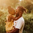 Esta imagem do Dia dos Pais mostra um pai e filha de pele escura, abraçados ternamente. A menina, em um vestido amarelo, beija o nariz do pai, que tem dreadlocks. A luz dourada do sol poente realça a cena afetuosa em um ambiente natural.