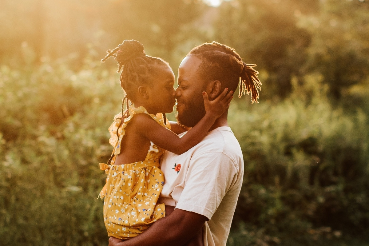Esta imagem do Dia dos Pais mostra um pai e filha de pele escura, abraçados ternamente. A menina, em um vestido amarelo, beija o nariz do pai, que tem dreadlocks. A luz dourada do sol poente realça a cena afetuosa em um ambiente natural.