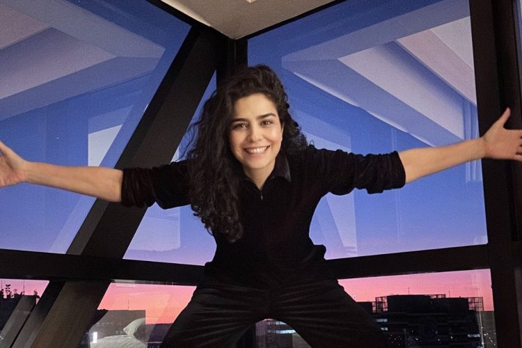 Leticia Sabatella de look preto, sorrindo, de braços abertos, com grandes janelas ao fundo
