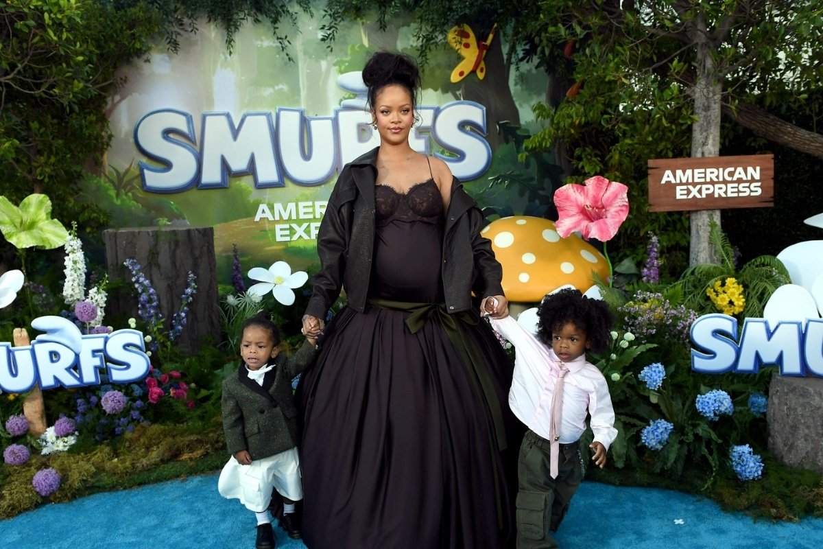 Rihanna na estreia de Smurfs em LA usando vestido preto com busto rendado, laço verde-oliva e jaqueta marrom segurando a mão dos filhos