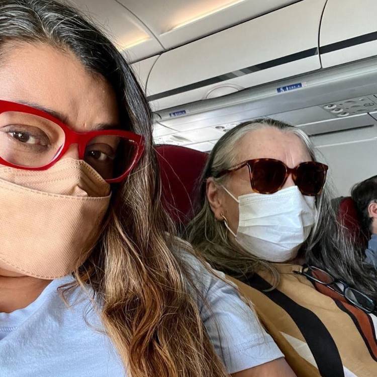 Duas mulheres, possivelmente mãe e filha, estão sentadas em um avião. A mulher da frente, à esquerda, usa óculos vermelhos e uma máscara bege. A mulher de trás, à direita, usa óculos escuros e uma máscara branca, com cabelos grisalhos.