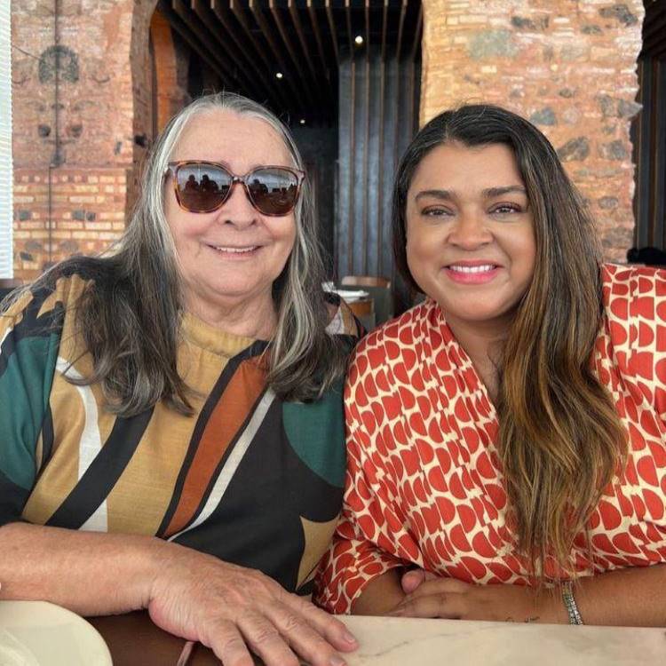 Duas mulheres, possivelmente mãe e filha, sorriem para a câmera em um ambiente interno. A mulher à esquerda tem cabelos grisalhos e usa óculos escuros e uma blusa estampada em tons terrosos. A mulher à direita tem cabelos castanhos e usa uma blusa estampada em vermelho e branco.