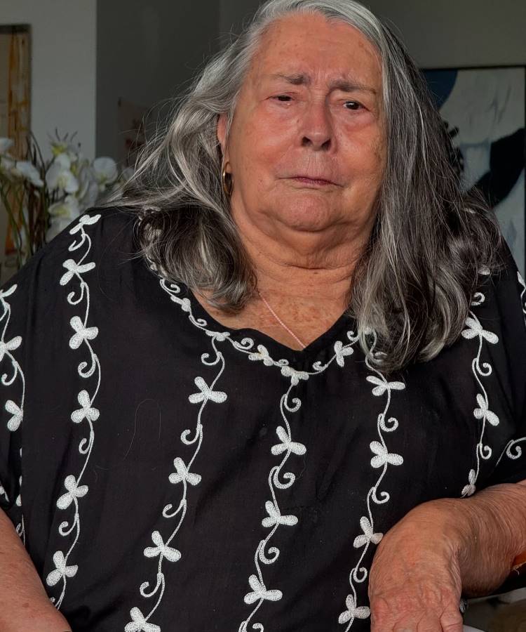 Uma mulher de idade, com cabelos grisalhos e longos, está sentada e olha diretamente para a câmera. Ela veste uma blusa preta com bordados brancos em formato de folhas e tem brincos dourados. Ao fundo, há um arranjo de flores brancas.
