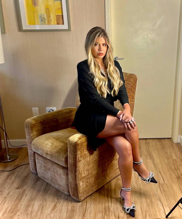 Cantora Marília Mendonça que faria 30 anos em 2025 de vestido preto curto sentada no braço de poltrona caramelo, em local interior.
