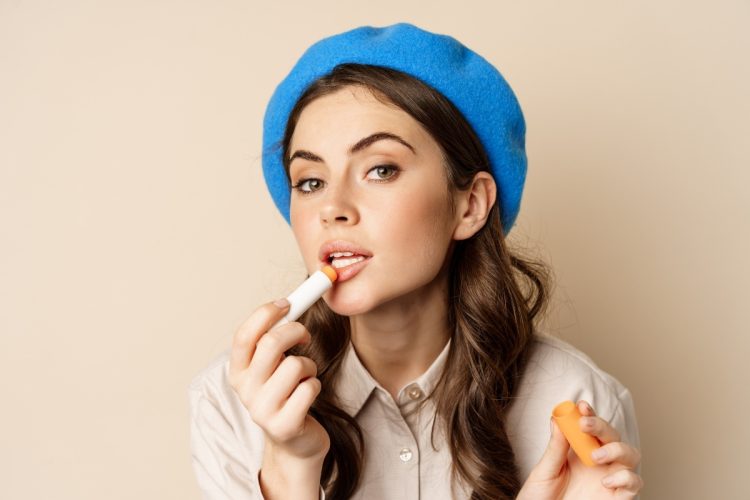 Mulher de pele clara usando roupa bege, boina azul, passando hidratante labial