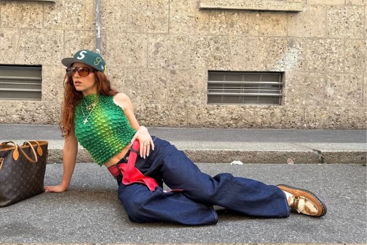 Mulher de pele clara deitada na rua usando blusa verde, calça jeans, mocassim cow print de camurça e boné