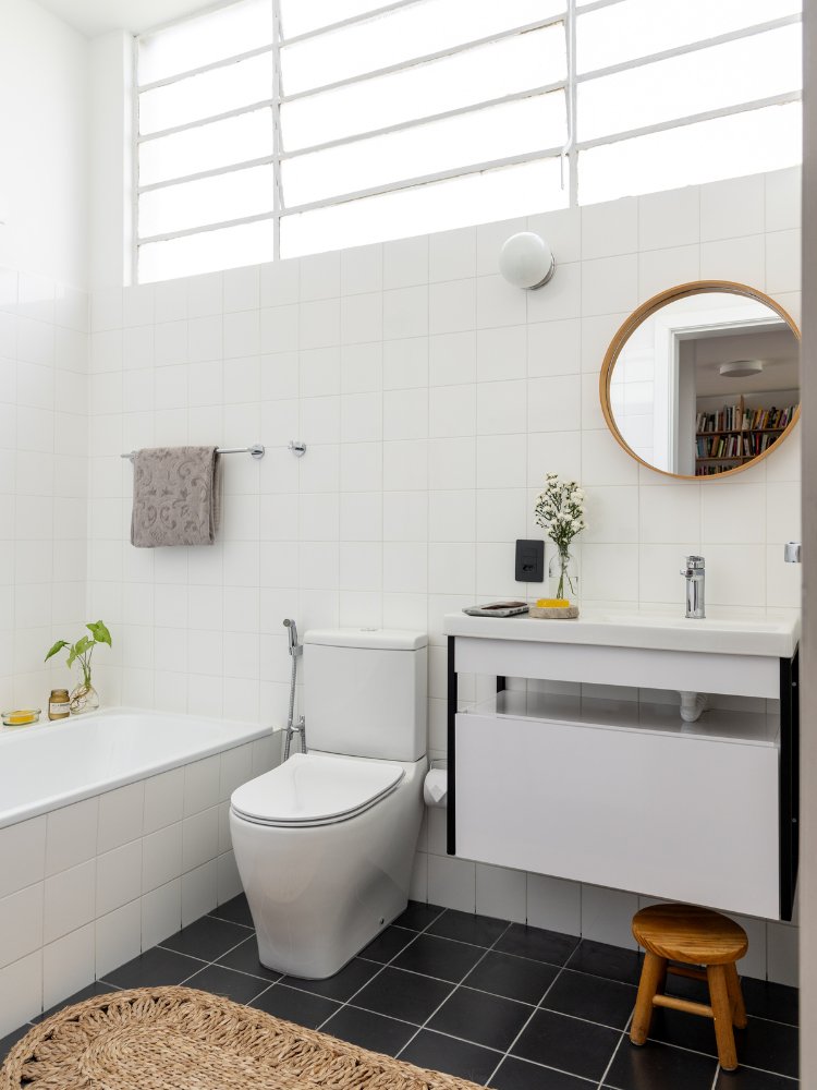 Visão mais geral do banheiro, com banheira, vaso sanitário, pia, gabinete e espelho redondo 