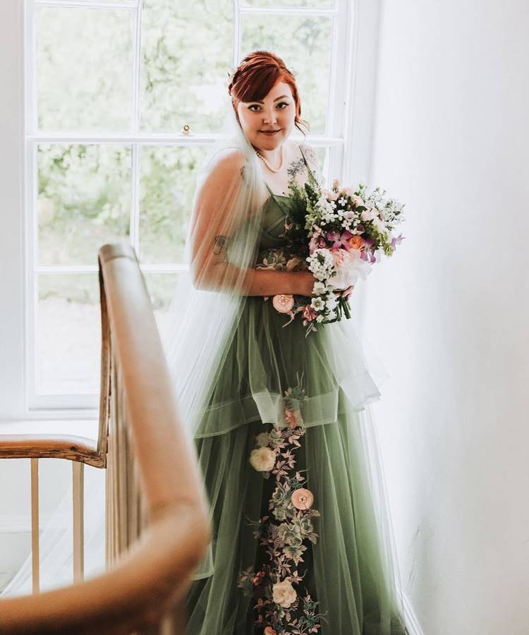 Uma noiva sorridente com cabelo ruivo preso e véu curto, usando um vestido de tule verde-oliva em camadas, com bordados florais rosados, posa segurando um buquê colorido ao lado de uma escada e janela.