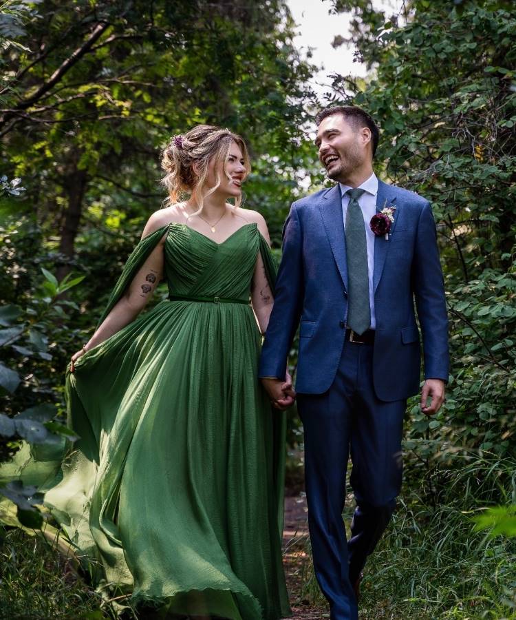 Um casal de noivos de mãos dadas, caminhando por um caminho arborizado. A noiva sorridente usa um vestido verde-oliva esvoaçante e o noivo veste um terno azul com gravata verde e flor na lapela.