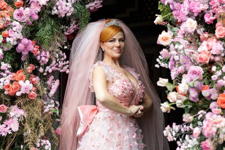 Uma noiva sorridente com cabelo ruivo e tiara, usando um vestido de noiva rosa claro com detalhes florais e um véu rosa. Ela está emoldurada por uma exuberante decoração floral em tons de rosa, branco e salmão.