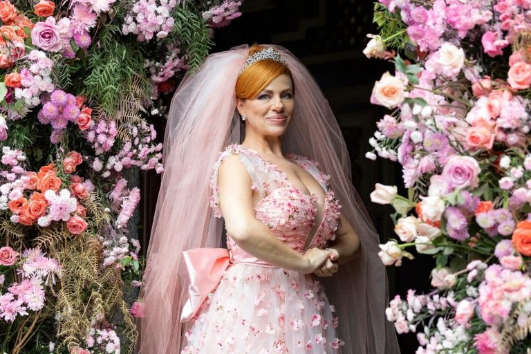 Uma noiva sorridente com cabelo ruivo e tiara, usando um vestido de noiva rosa claro com detalhes florais e um véu rosa. Ela está emoldurada por uma exuberante decoração floral em tons de rosa, branco e salmão.