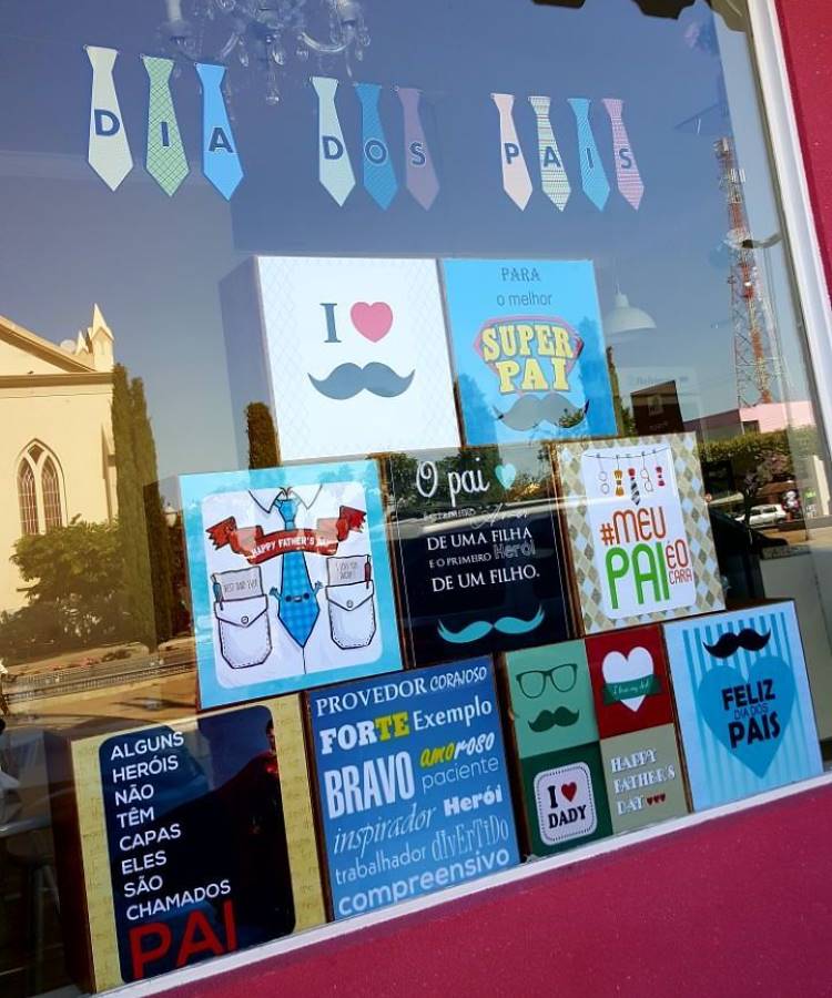 A imagem exibe uma vitrine decorada para o Dia dos Pais. Na parte superior, gravatas de papel formam "DIA DOS PAIS". Abaixo, diversos quadros e caixas com mensagens e ilustrações temáticas de pai, incluindo bigodes, frases carinhosas e homenagens.
