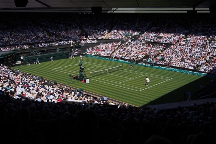 Foto de partida de tênis no torneio de Wimbledon, com tenistas e espectadores usando roupas brancas
